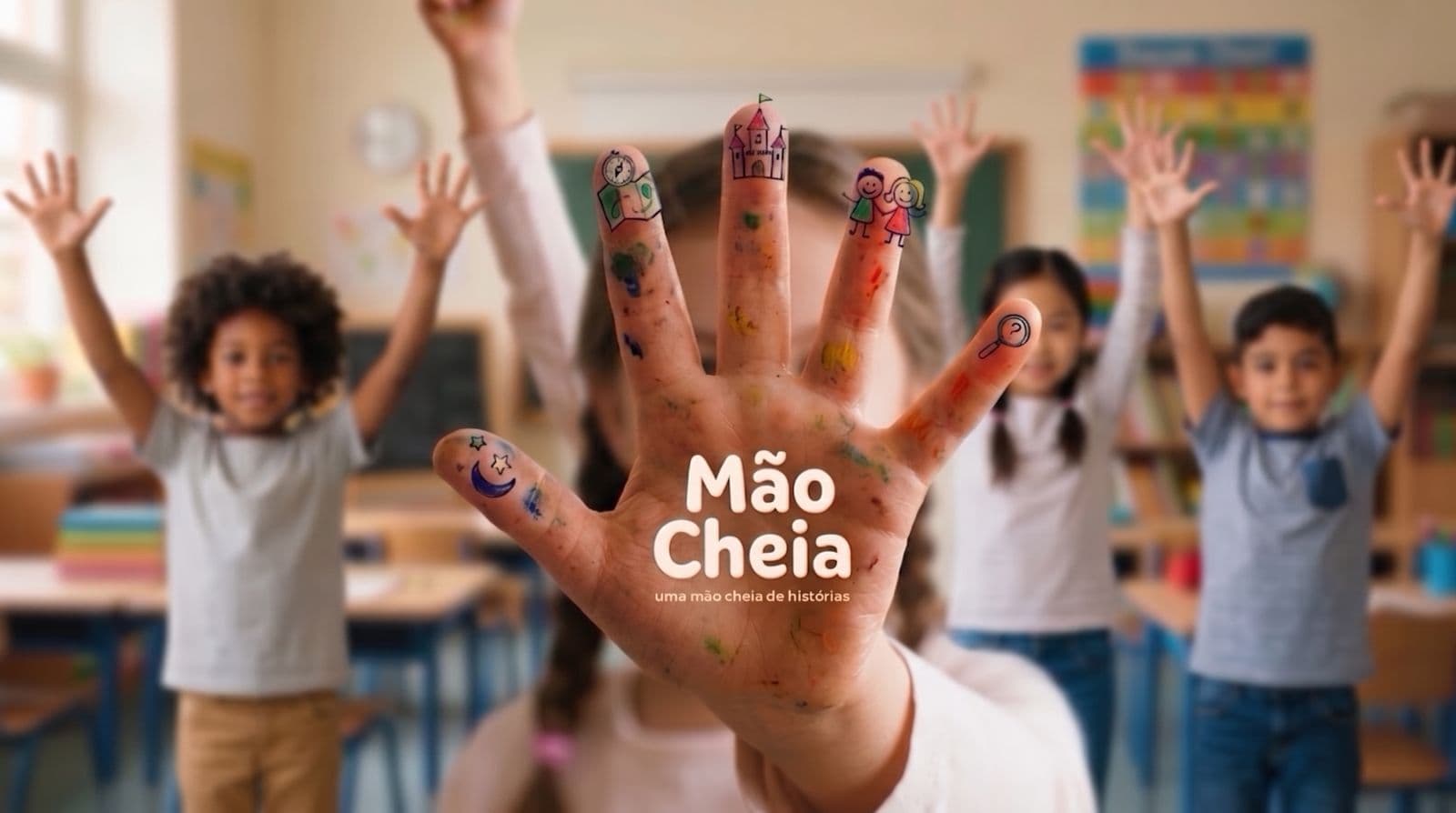 A mão aberta de uma criança em primeiro plano, suja de tinta, com pequenos desenhos pintados nas pontas dos dedos e o nome «Mão Cheia — uma mão cheia de histórias» impresso na palma; ao fundo, desfocada, uma sala de aula com três crianças de pé, de braços no ar.